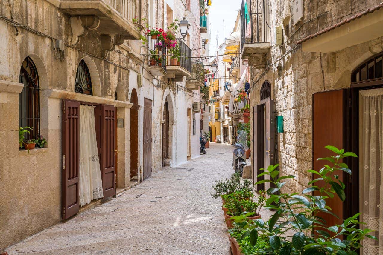 Una via stretta in pietra rosata con diverse porte e balconi decorati con vasi di piante