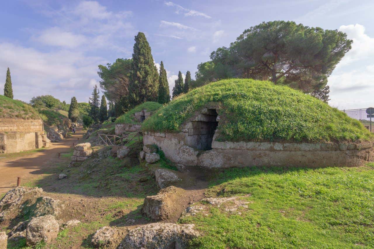 Ancient structire covered in grass, with stone entrances partially exposed. The scene is set against a bright, sunny sky with trees and shrubs scattered in the background.