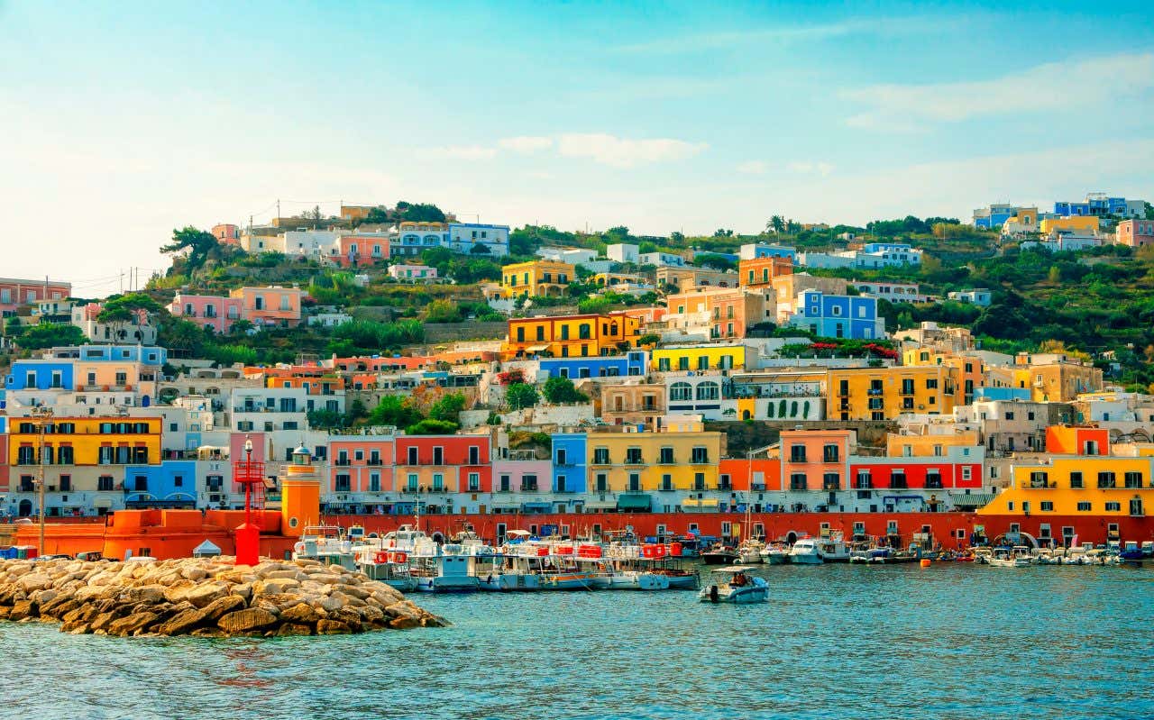 A hillside filled with colorful buildings in shades of blue, yellow, orange, and pink overlooks a marina with boats anchored near a rocky pier and a small red lighthouse.