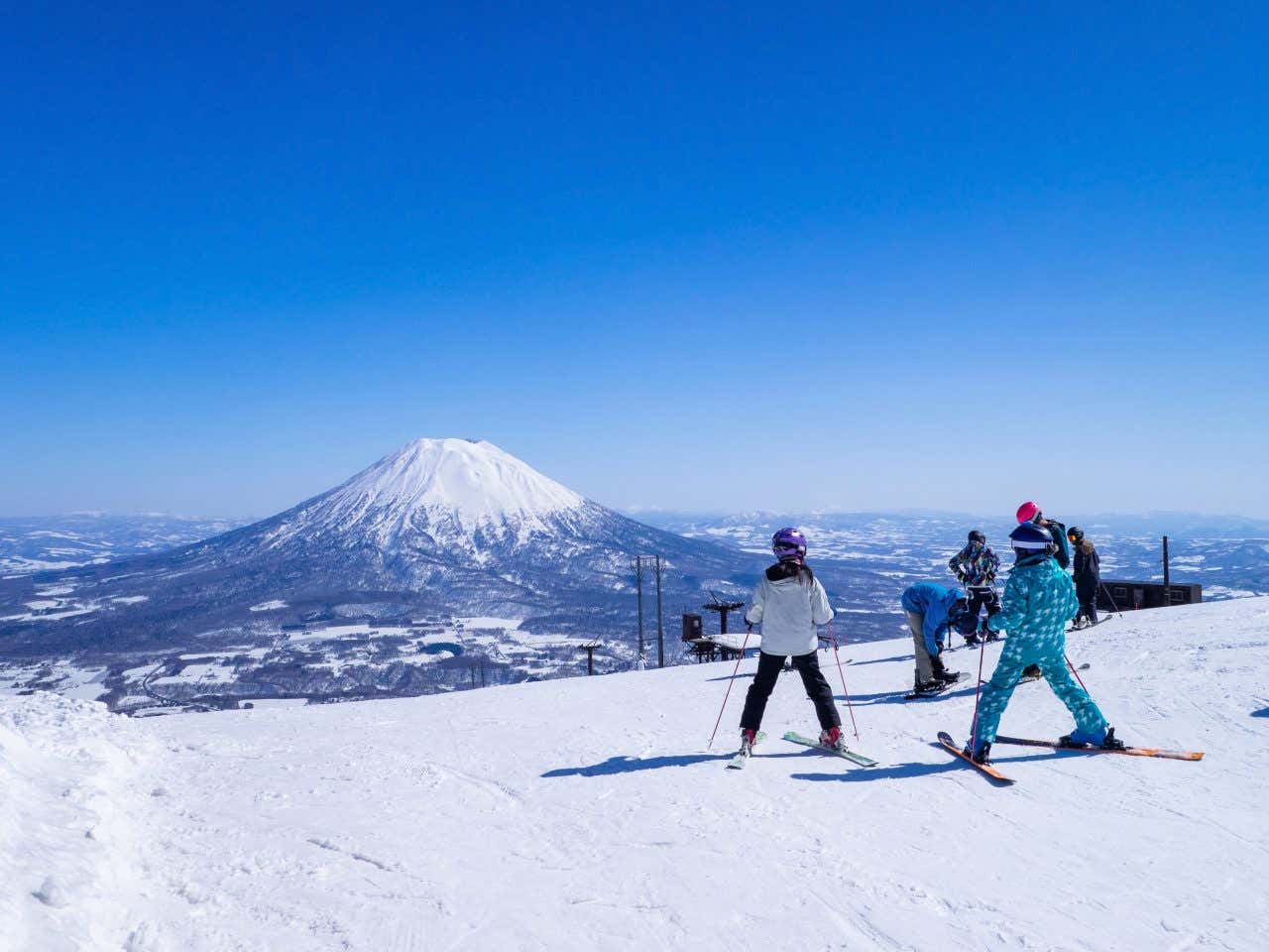 Various people locked into skis stand at the top of a run, with the Mount Yōtei clearly visible in the distance, from the Niseko ski resort, a top ski destination.