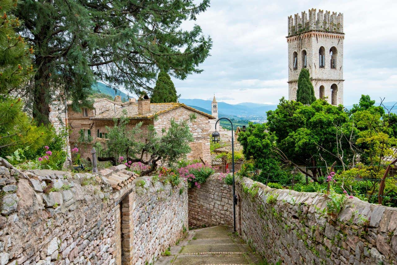 A stone pathway lined with flowers and greenery leads to a historic village with stone buildings and tall bell towers, surrounded by trees and distant hills.