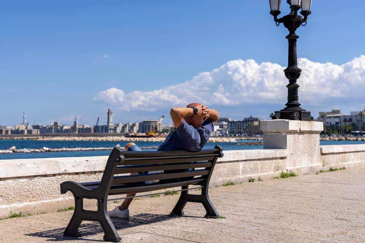 Un uomo seduto su una panchina del lungomare di Bari che si gode il sole, sullo sfondo un lampione nero in ghisa e il porto