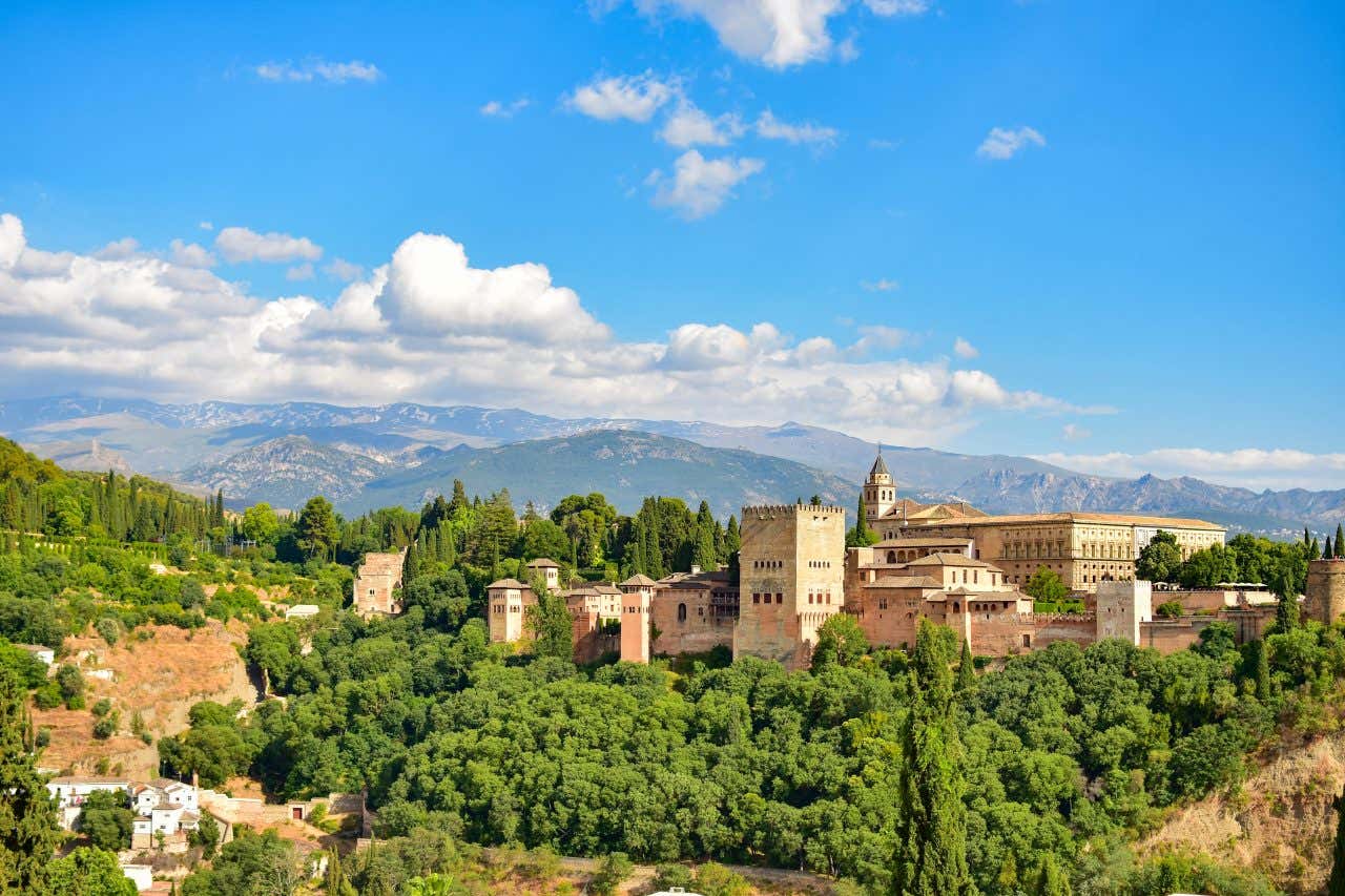 The Alhambra in Granada as seen from afar, with a mountain range, and a cloudy blue sky visible in the background.