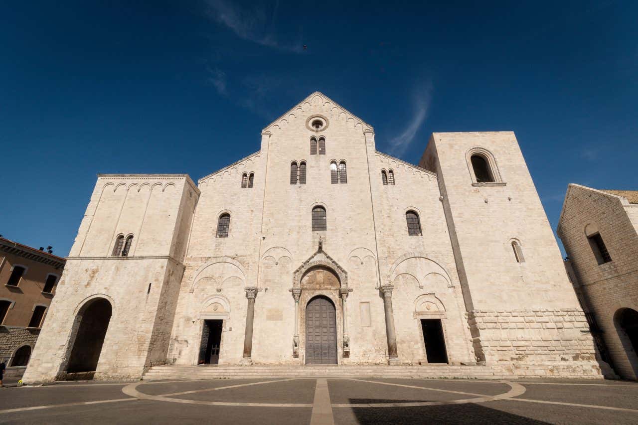 Facciata di una chiesa romanica in pietra bianca con due campanari laterali e una facciata centrale più alta, sormontata da un piccolo rosone in una giornata di sole senza nuvole