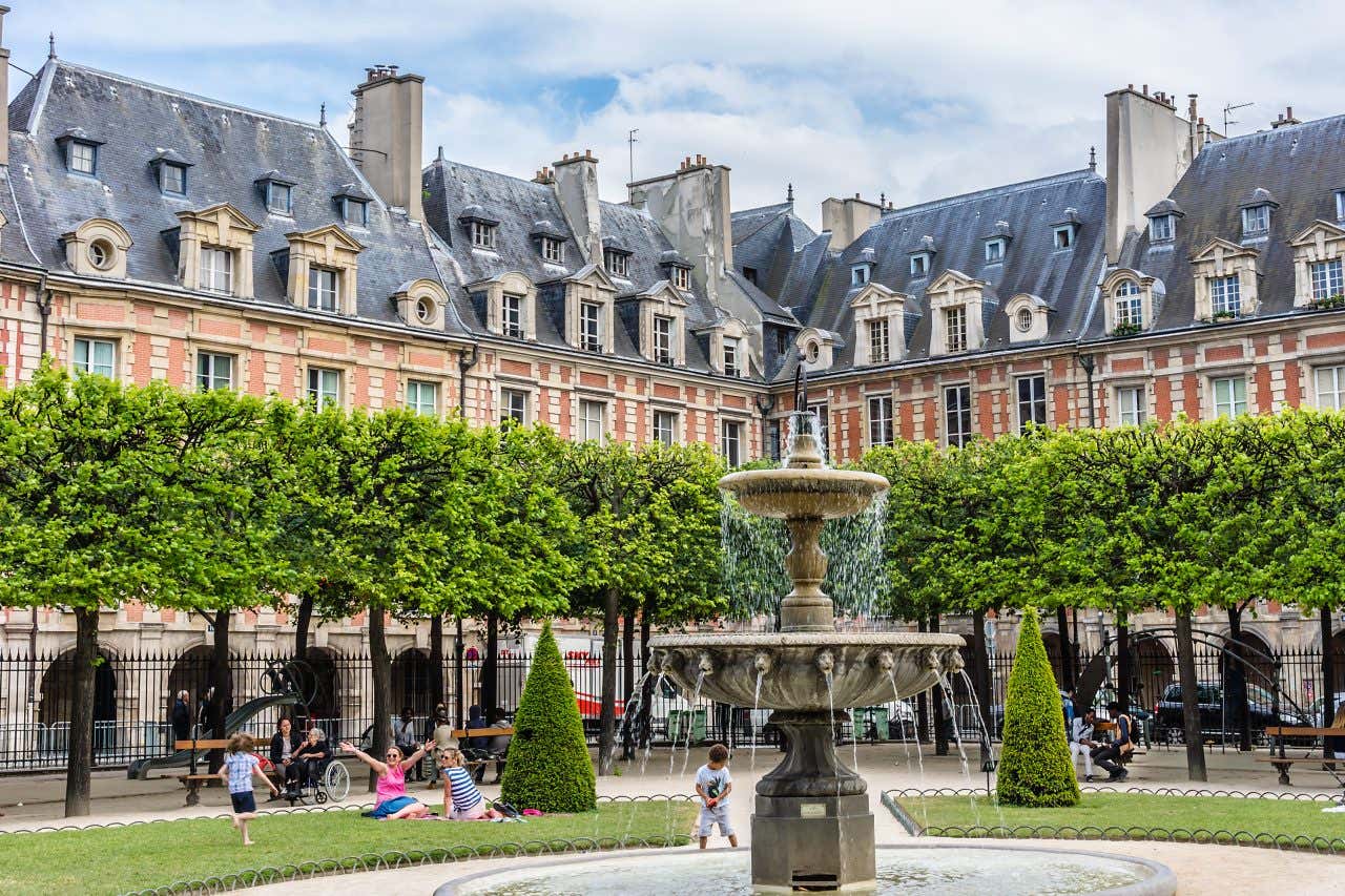 A square  in Paris with buildings in the background, and green trees in the foreground, as well as a water fountain.