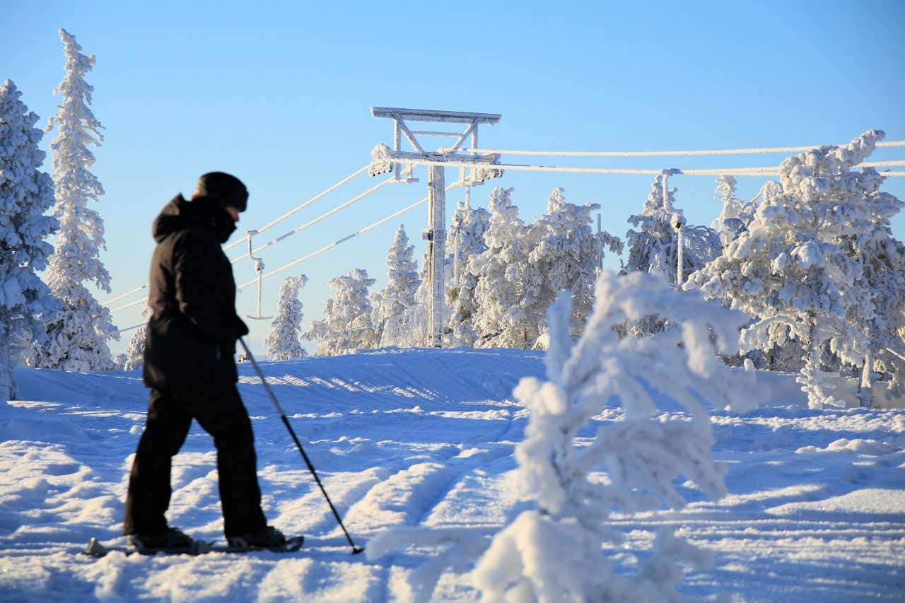 A lone skiier dressed all in black walks across an emtpy ski run, a ski lift visible behind covered in snow, within the FInnish laplands.