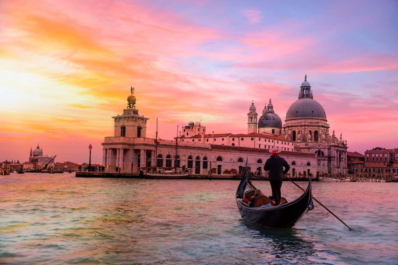 Une gondole sur l'eau avec une personne debout la dirigeant et la basilique en fond sous un ciel aux tons rosés