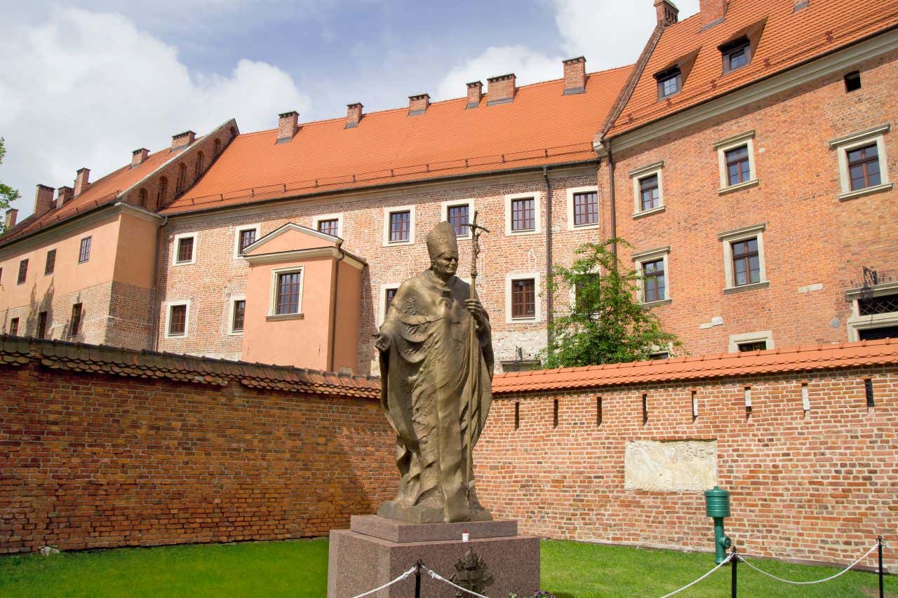 La statua del papa in bronco al centro di un cortile urbano circondato da case in mattoni rossi in un giorno di sole