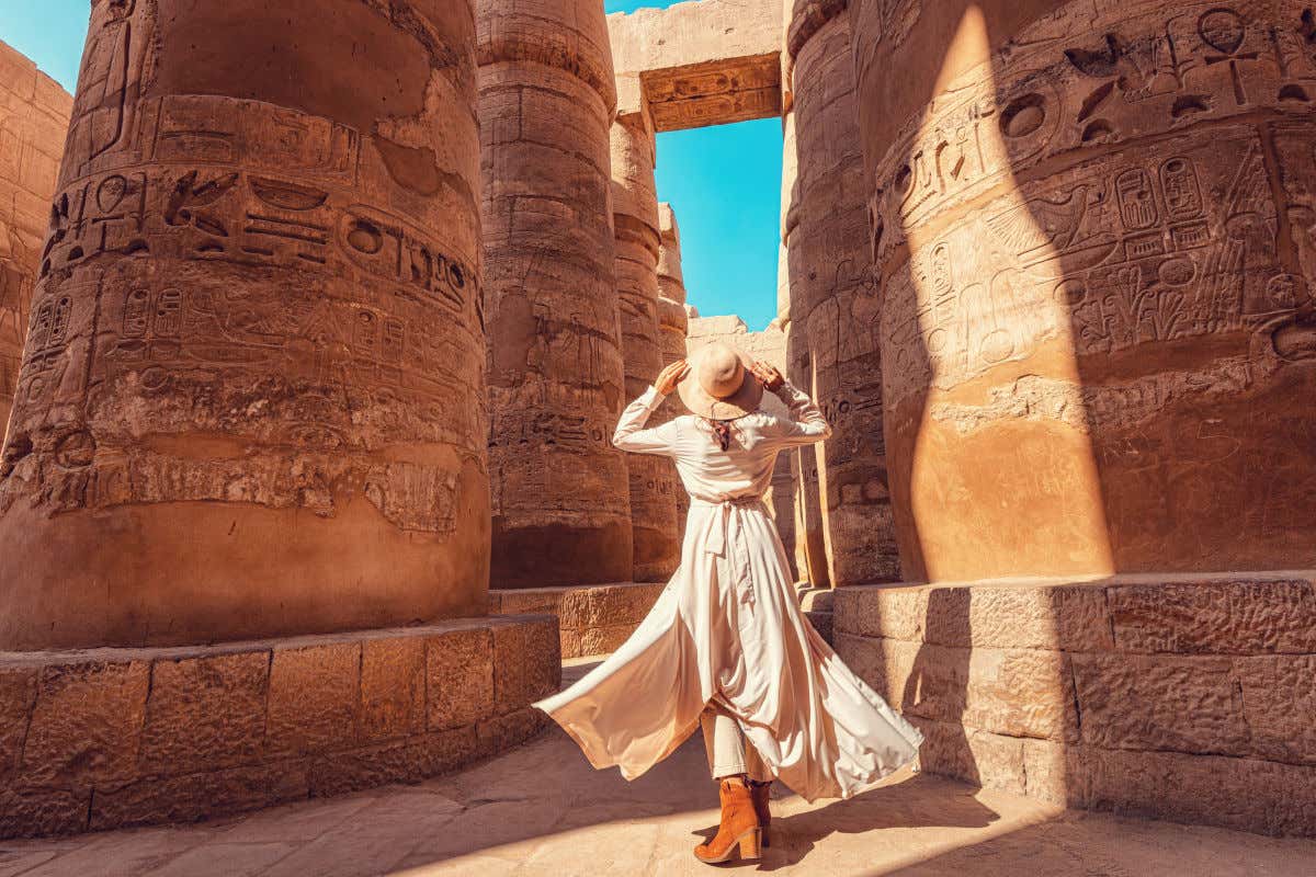 A woman with her back to the camera wearing a dress and hat gazing at the tall columns of the Karnak temple in Egypt.