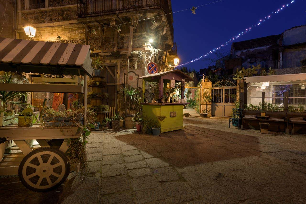 Un angolo di strada urbano fotografato di sera, circondato da palazzi storici, illuminato cn lampioni e lucine e decorato con carretti di legno su cui sono poggiati piccoli vasi di piante