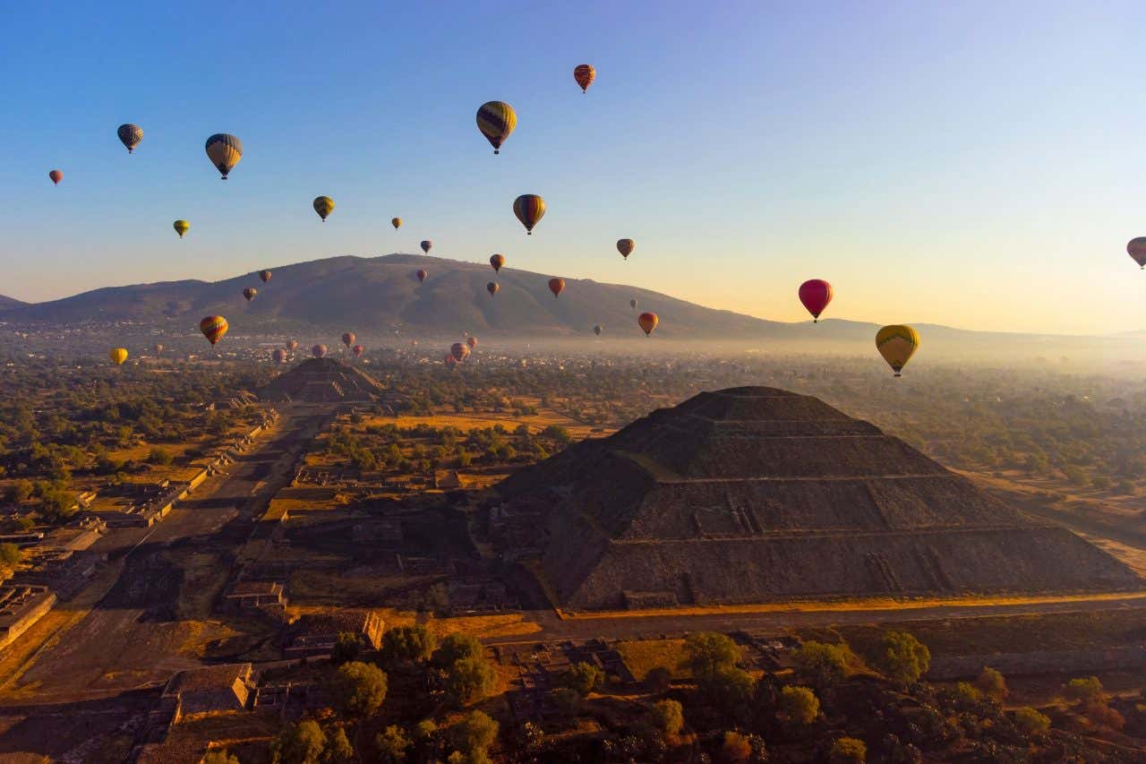 The Pyramid of the Sun as seen from the air, with hot air balloons flying in the background. and a blue sky in the background with some yellow at the bottom.