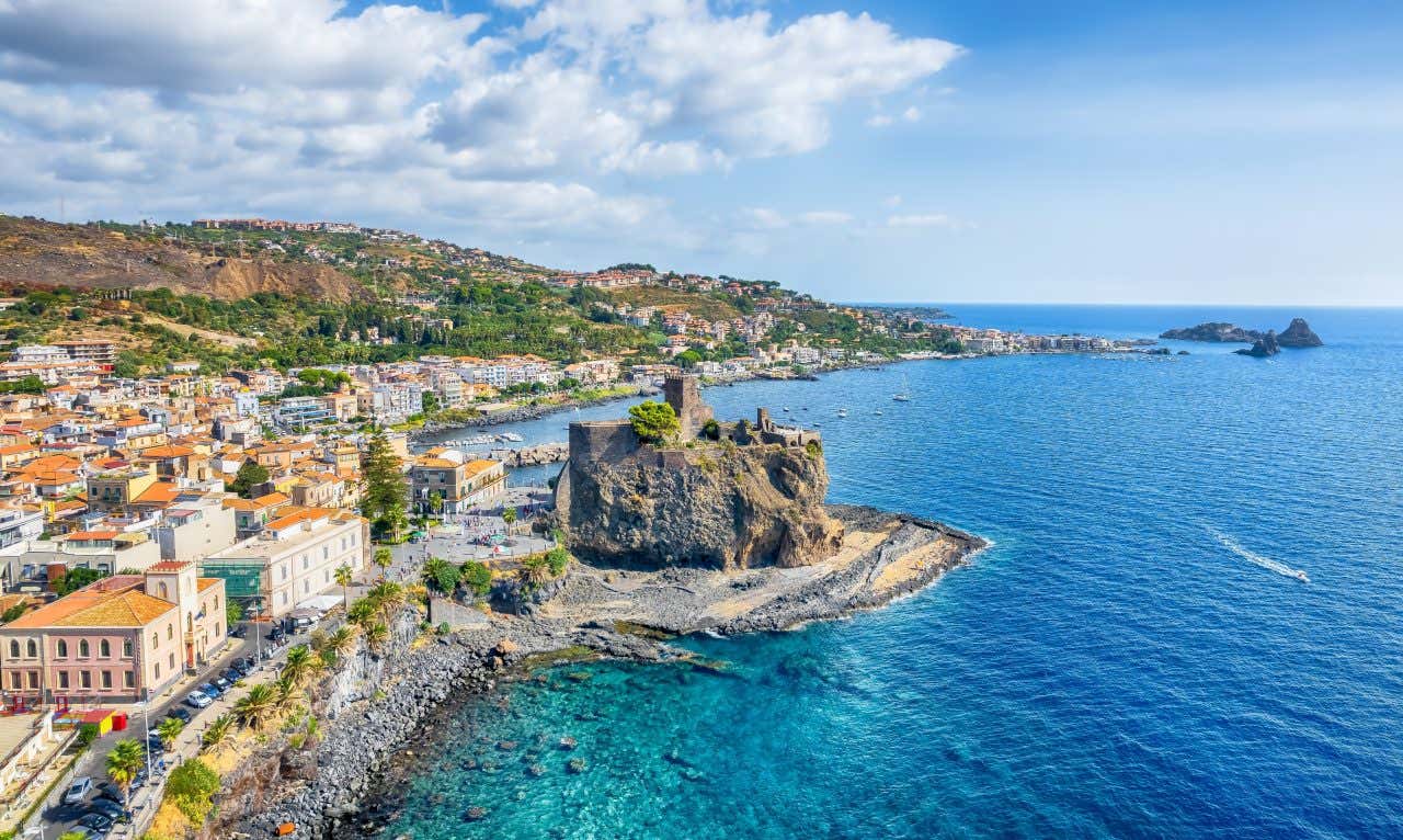 Veduta aerea della costa di Aci Castello, con il Castello Normanno arroccato su una scogliera di roccia lavica che si affaccia sul mare. Le acque cristalline circondano la fortezza, mentre le case del borgo si distribuiscono lungo la costa, creando un suggestivo panorama mediterraneo