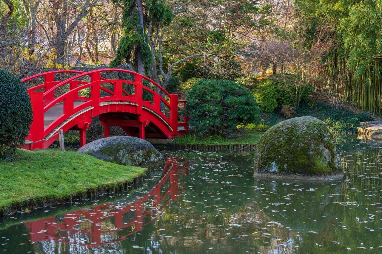 Jardin Pierre Baudis de style japonais, une visite à faire à Toulouse ; avec un pont rouge à gauche, un plan d'eau et deux rochers dans l'eau