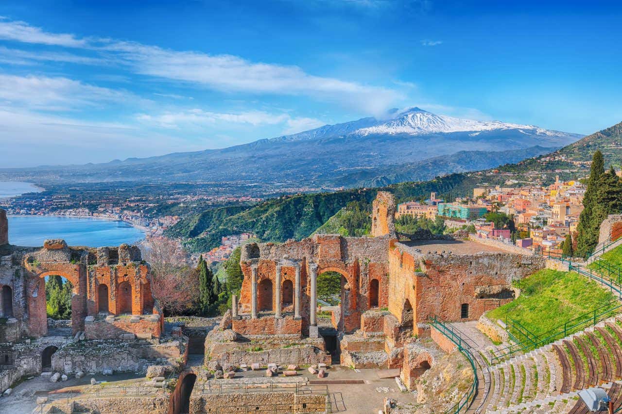 Resti archeologici di Taormina, con le antiche colonne e le gradinate del Teatro Greco in primo piano. Sullo sfondo, l’Etna innevato si staglia maestoso contro il cielo, creando un contrasto suggestivo 