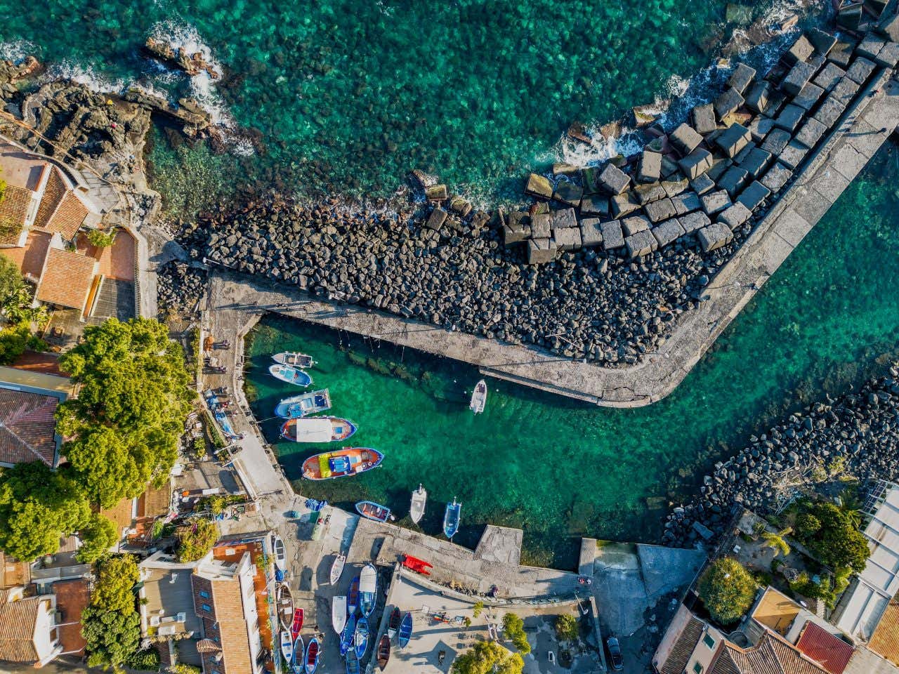 Un piccolo porto con il molo in cemento in mezzo al mare turchese e trasparente, con alcune barchette in legno attraccate nei pressi di piccole case dal tetto giallo