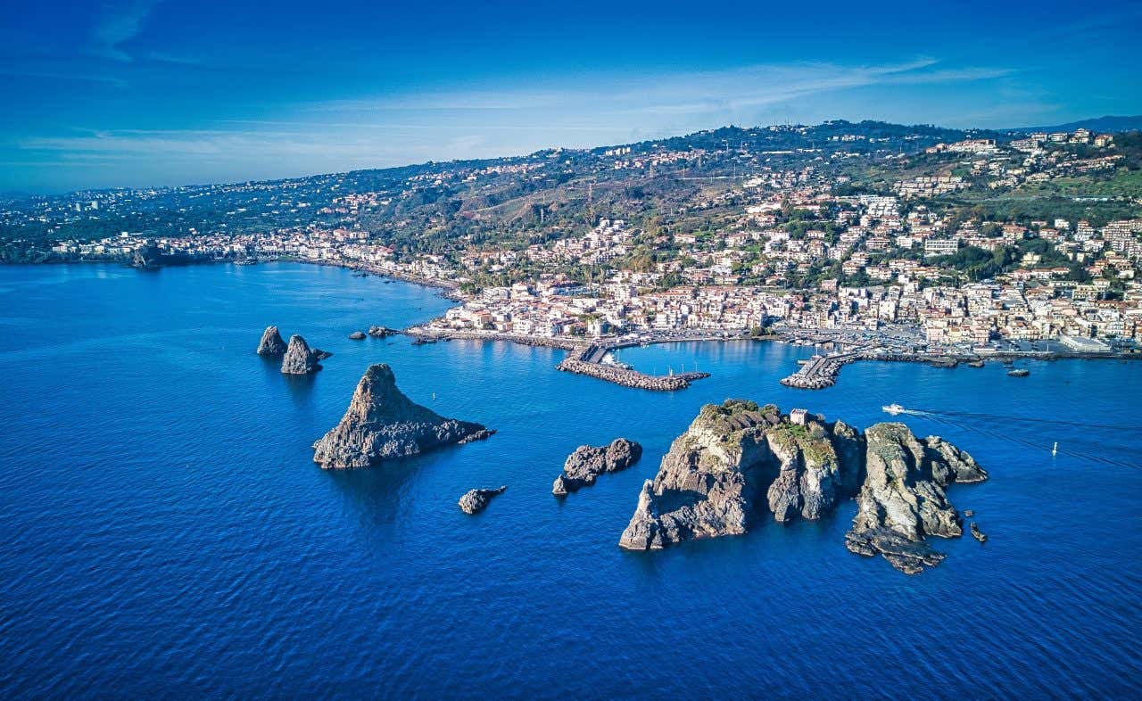 Veduta aerea della costa di Aci Trezza, con le Isole dei Ciclopi che emergono dalle acque turchesi del Mar Ionio. I faraglioni di origine vulcanica creano un paesaggio mozzafiato, mentre il borgo marinaro si sviluppa lungo la costa, con i moli del porto che si estendono sull'acqua