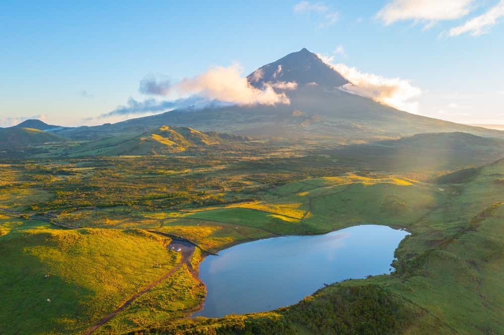 Una vallata fotografata alle prime luci dell'alba, un lago in primo piano circondato da colline verdi, in fondo un monte coperta da nubi