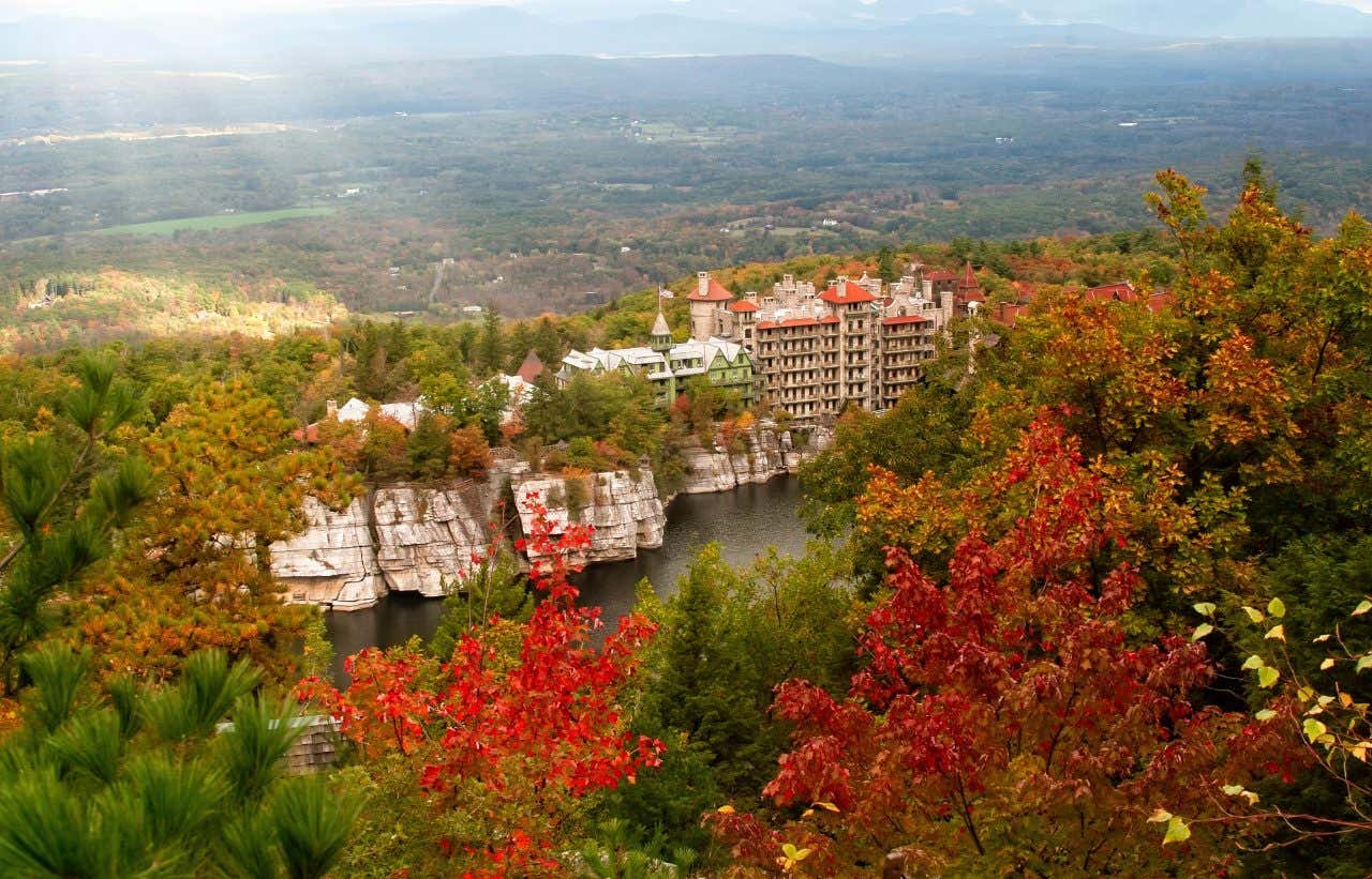 An aerial shot of Mohonk Preserve in New York during the fall, with trees with red and orange leaves in the foreground, and a large building across the water.