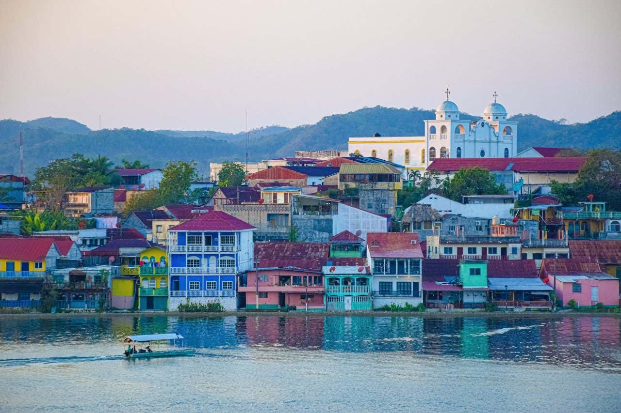 A shot of Flores, Guatemala as seen from across a body of water, with a boat sailing from left to right. 