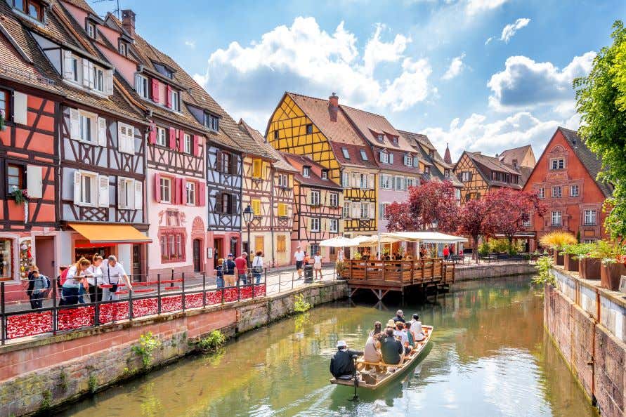 A canal in Colmar with people on a boat along it and colorful houses lining the canal under a cloudy sky.