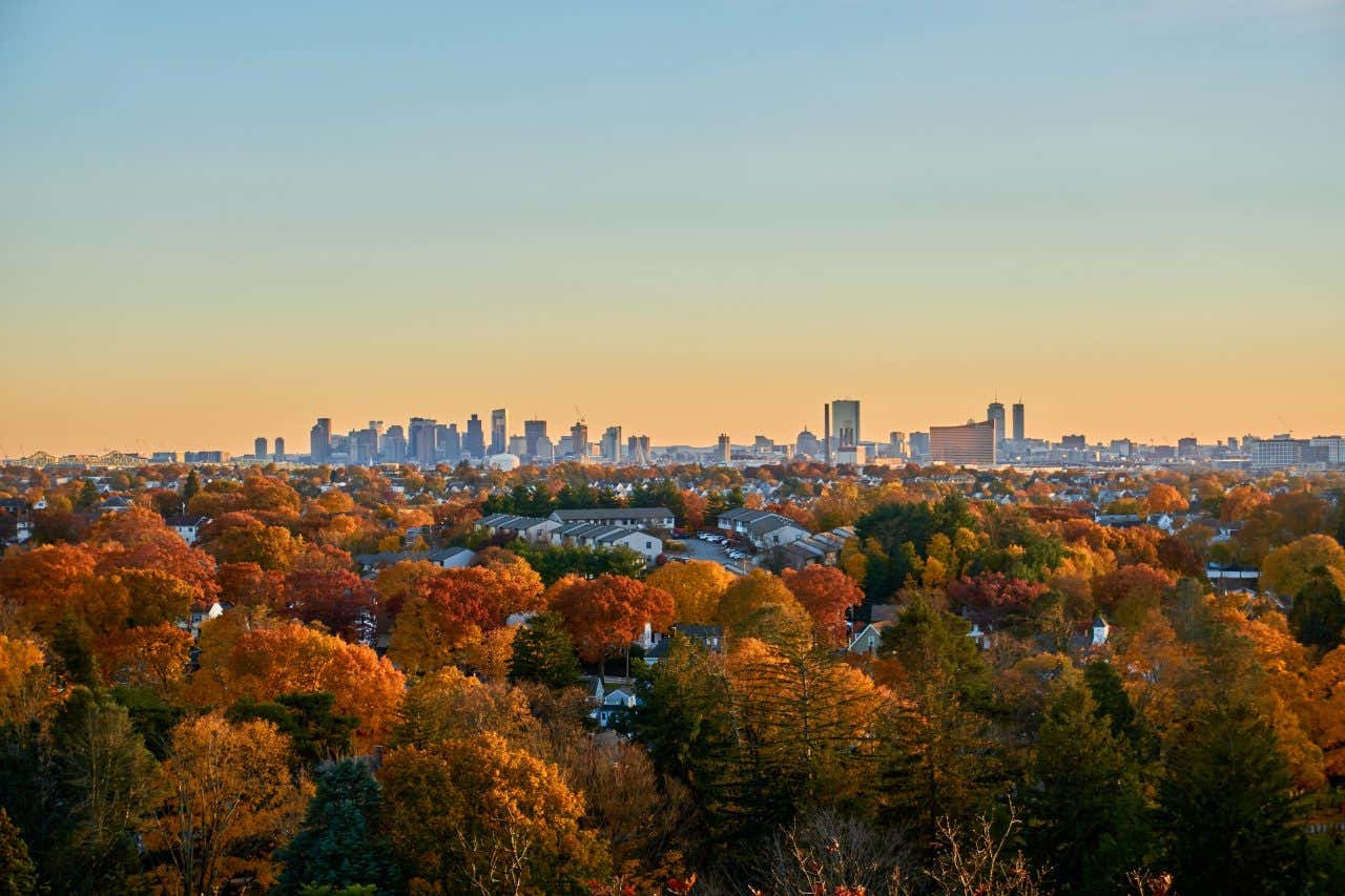 Boston as seen from afar, with buildings in the skyline, and blue and orange skies.