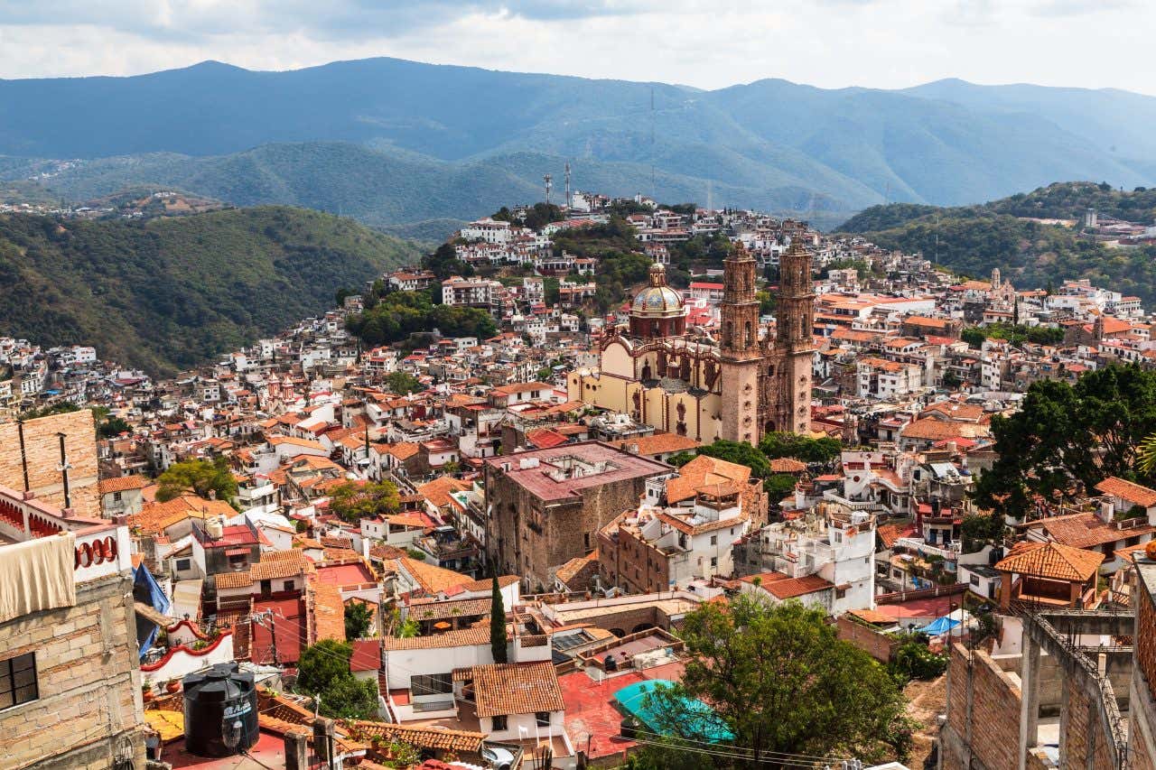 Taxco as seen from above, lots of colorful buildings take up the bottom half of the picture, and mountains are visible inthe background.