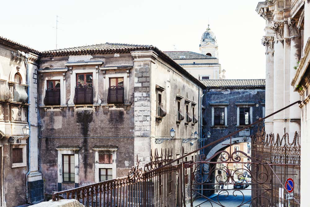 Palazzi antichi in pietra nera e bianca nel centro di Catania, sullo sfondo un arco da cui si intravedono palazzi più recenti