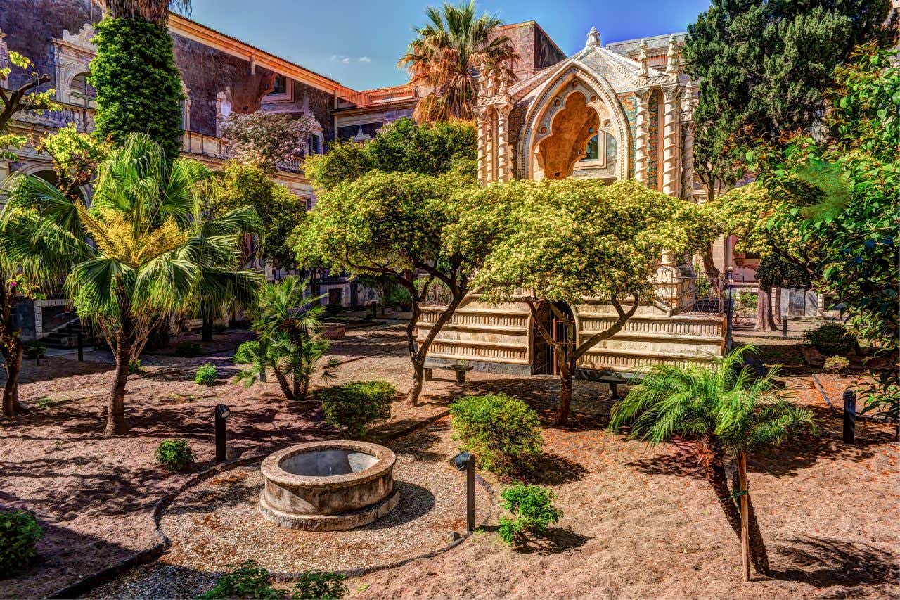 Chiostro del Monastero Benedettino di San Nicolò l'Arena a Catania, con eleganti colonne in pietra lavica e un giardino centrale verde, incorniciato da archi barocchi. La luce naturale illumina l'architettura storica, evidenziando i dettagli decorativi