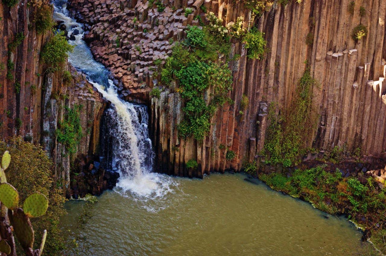 A waterfall leadign in to a body of brown water, surrounded by tall cliffs.