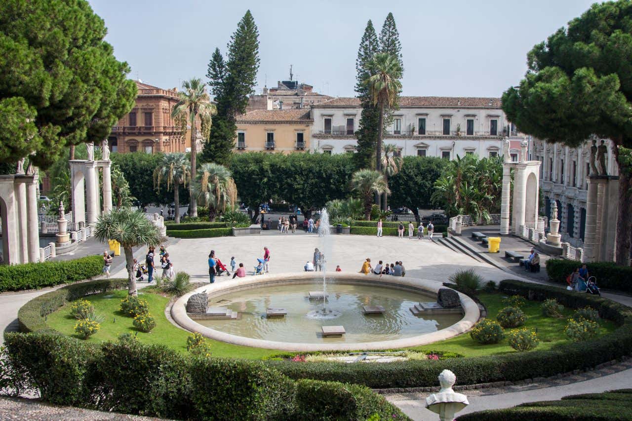 Diverse persone sedute sul bordo di una fontana circolare al centro di un giardino ben curato, in cui prati e aiuole si alternano a scalinate, colonnati e statue