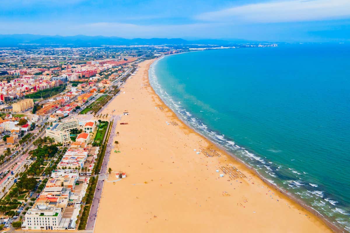 Vista aérea de una playa, de fina arena dorada y aguas de azul turquesa junto a una hilera de edificios