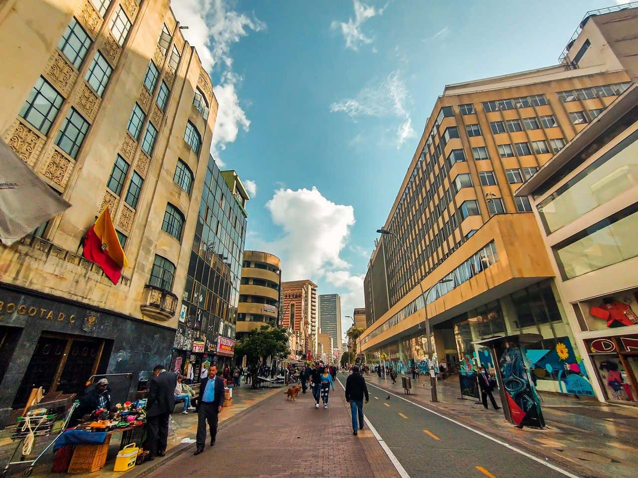 A street in Bogotá, Colombia, with people walking up and down the street, and a cloudy blue sky in the background.