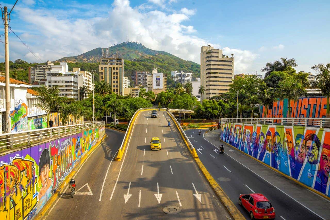 A street in Cali, Colombia with cars and motorbikes driving each way, colorful grafitti on walls on either side of the road and a green mountain in the background.